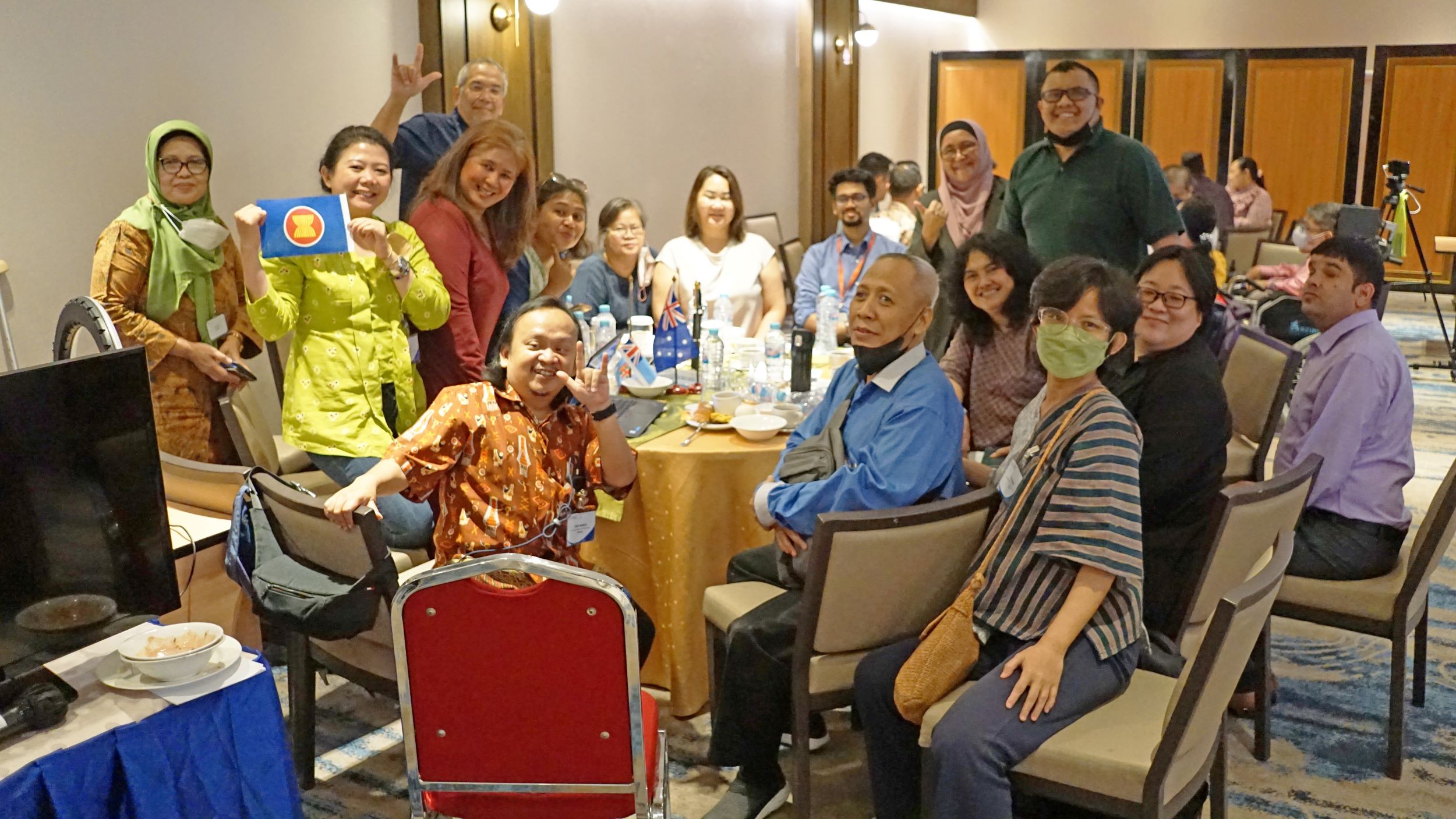 A group of more than a dozen people stand and sit around a table. The people have varying gender presentations and disabilities. Many of them are smiling and forming "love" signs with their hands. One is holding up a small ASEAN flag.