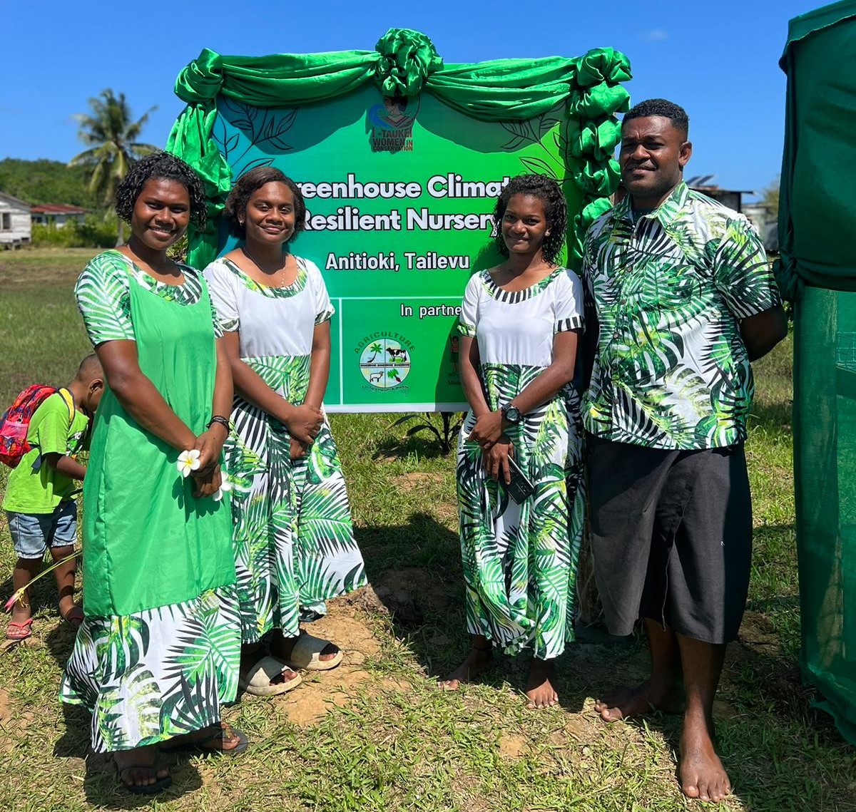 Four young brown people stand side-by-side and pose for the camera, smiling. They are wearing bright outfits with fern leaves on them. Behind them is a sign that reads "Greenhouse Climate Resilient Nursery - Antioki, Tailevu."