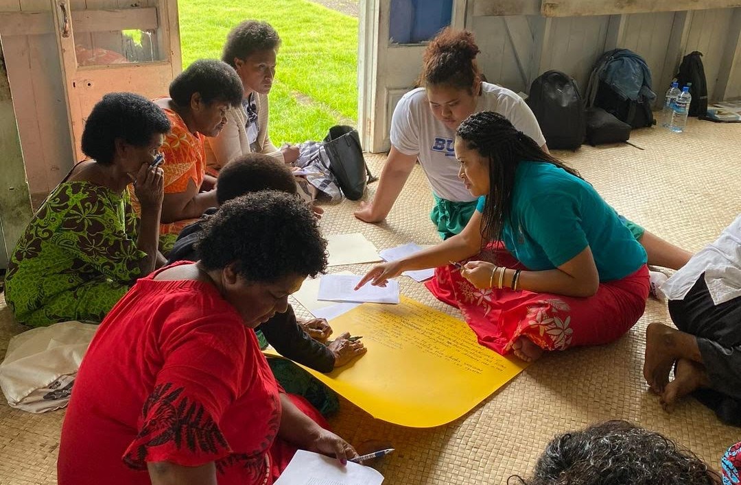 Several brown women sit in a circle on the floor of a room, many pieces of paper between them. They appear to be in the middle of discussion and writing notes on a large yellow piece of poster paper.