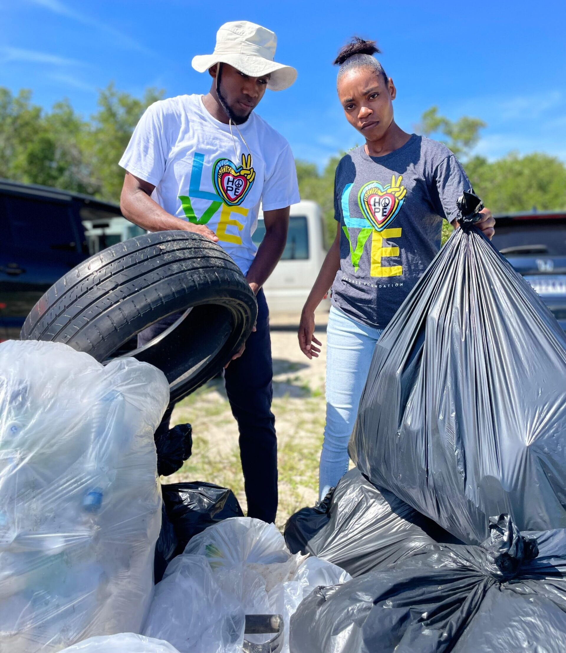 Two Black people stand next to each other, a pile of trash bags in front of them. The person on the left is holding up a tire, while the person on the right is holding up a trash bag and looking directly at the camera with a serious expression.