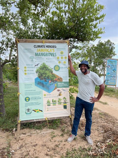 Mikhail Jobson stands on sandy ground that is full of plant life. He is facing the camera and smiling, posing next to a large sign with the heading "Climate Heroes: Jamaica's Mangroves" in bold text. Underneath the heading is an illustration of mangroves and lots of smaller text.