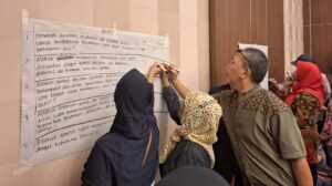 Three people with varying gender presentations face away from the camera and write on a large piece of paper that is taped to a wall. The text on the page is in Indonesian.