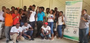 A group of young people from Haiti stand together as a group and pose for the camera. Most of them are holding their hand in one position, with their thumb, pointer, and pinky finger up. Many of them are laughing. Next to them is a banner that says in French: "Plaidoyer pour la reduction des effets du changement climatique sur les personnes handicapees." Beneath that text are logos for Global Greengrants Fund and our Haitian grantee partner CONAPH.