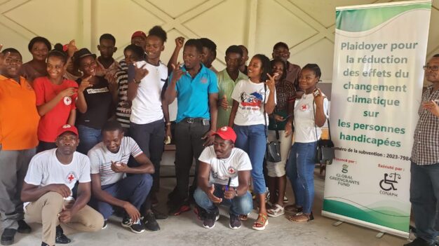 A group of young people from Haiti stand together as a group and pose for the camera. Most of them are holding their hand in one position, with their thumb, pointer, and pinky finger up. Many of them are laughing. Next to them is a banner that says in French: "Plaidoyer pour la reduction des effets du changement climatique sur les personnes handicapees." Beneath that text are logos for Global Greengrants Fund and our Haitian grantee partner CONAPH.