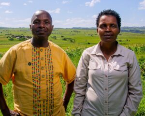 Sinegugu and Nonhle stand next to each other. Sinegugu is on the left. He is a Black man with a bald head. He is wearing a bright yellow shirt with colorful patterns embroidered onto it, and he is smiling slightly, his hands on his hips. On the right is Nonhle. She is a Black woman with curly hair cropped short. She is wearing a button up grey shirt and a more serious expression. Behind them is a lush and bright green field of grass.