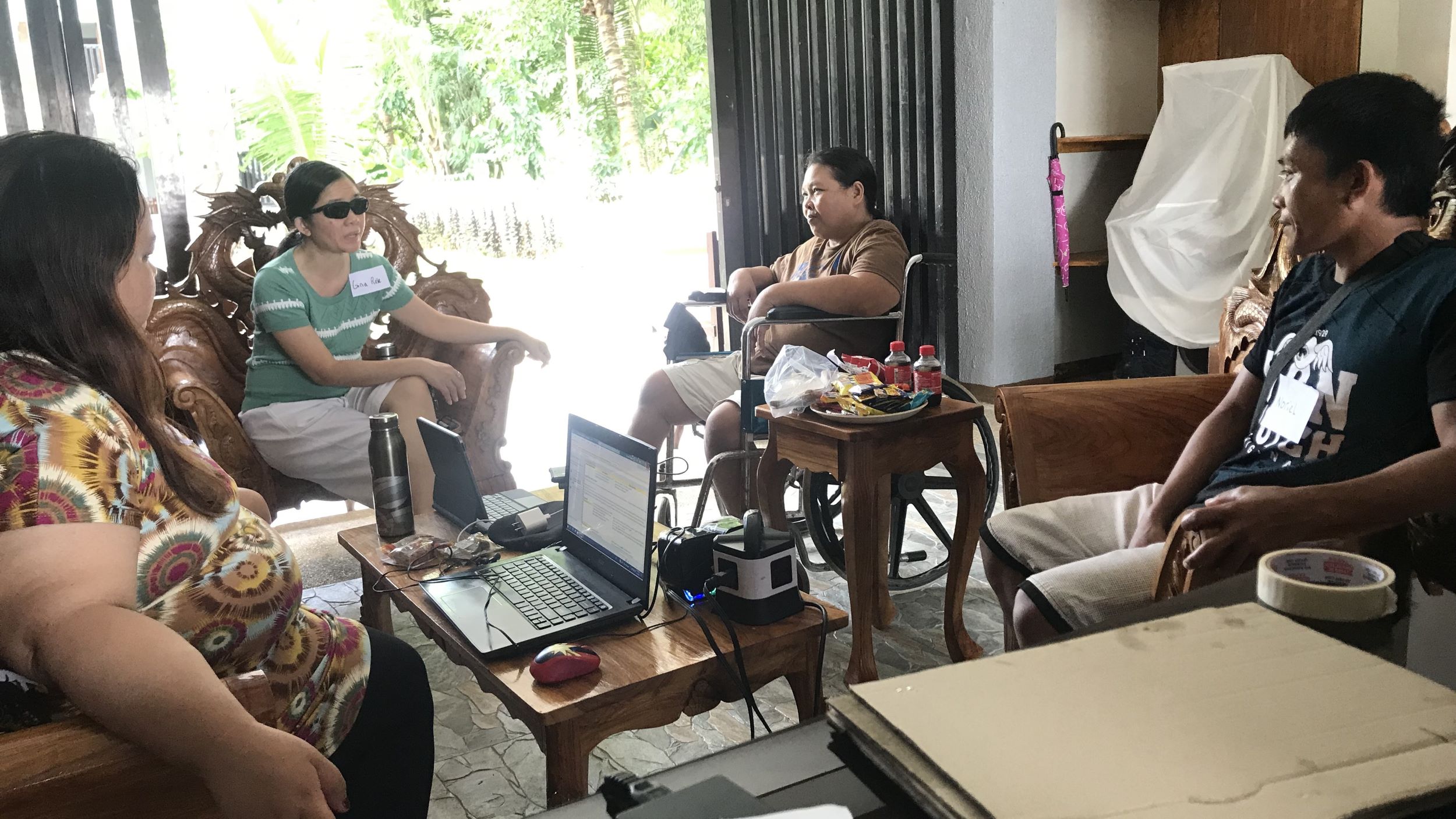 A group of people with varying disabilities and gender presentations sit around a table. The table has two open laptops and other electronic devices. They appear to be deep in conversation.