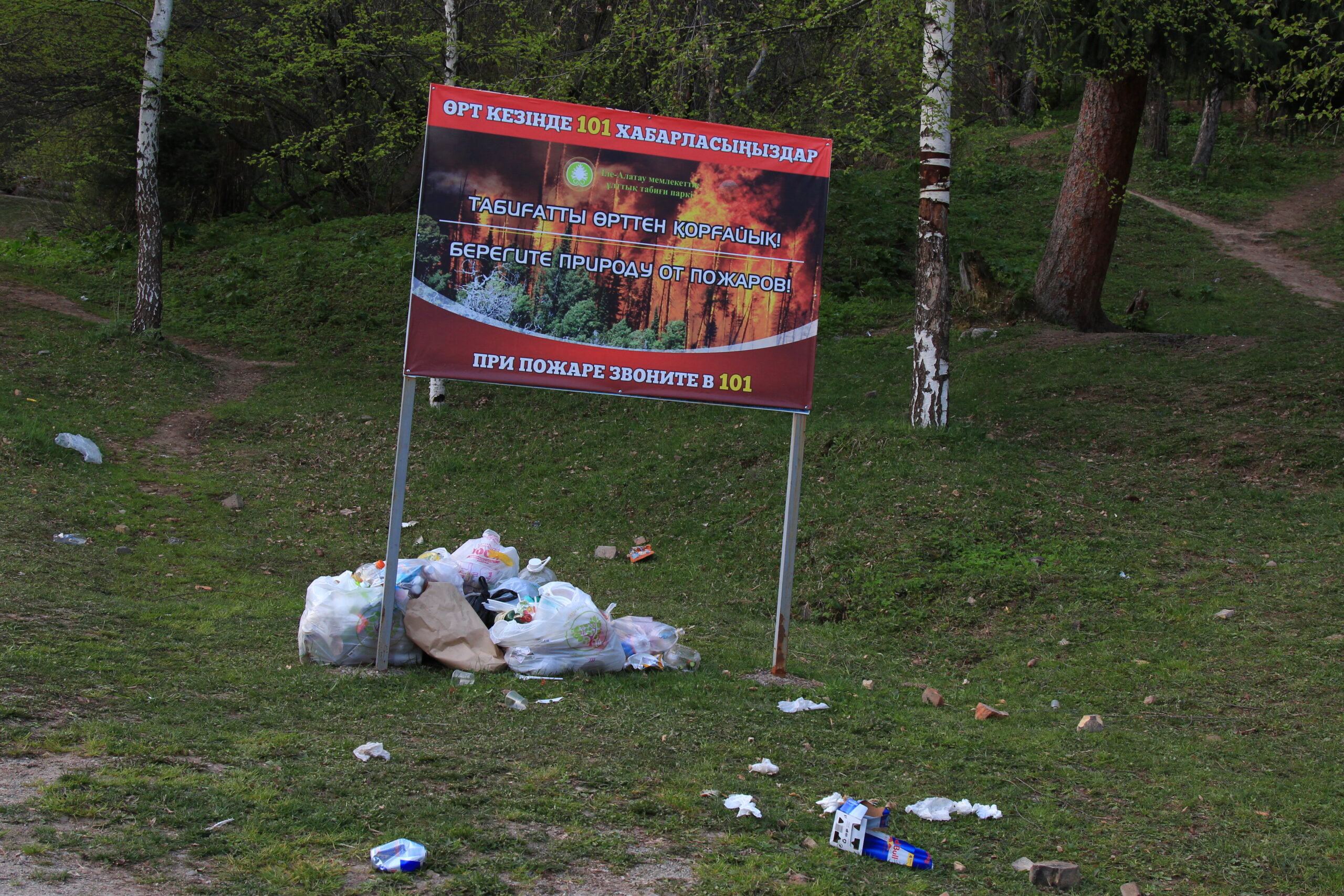 A sign featuring imagery of a forest fire sits on a hill covered in grass and trees. Under the sign are bags of trash, and there are various other pieces of trash scattered on the grass around the sign.