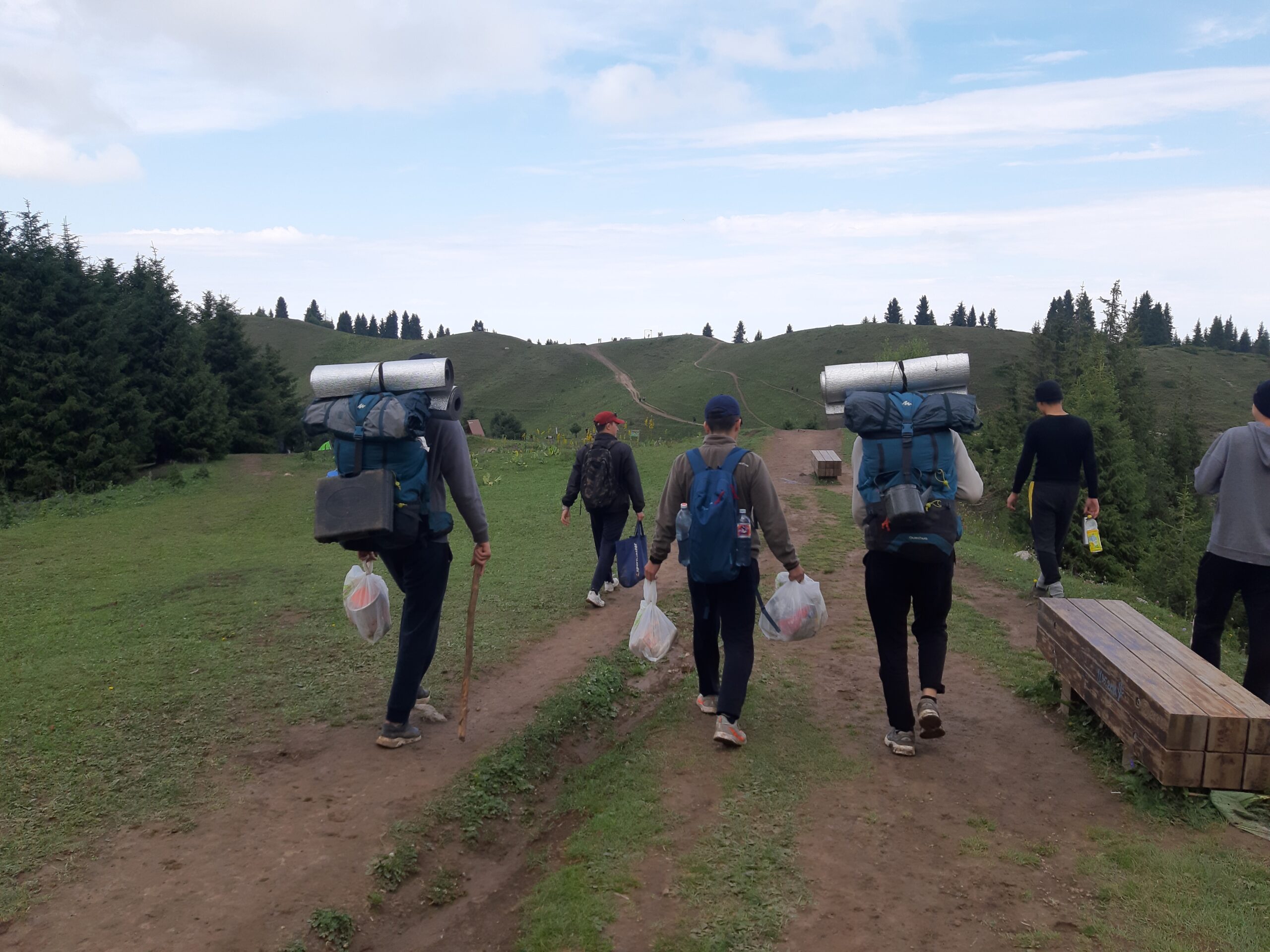 A group of people with backpacks and plastic bags walk up a hill, their backs to the camera. Around them, the hill is covered in grass and trees.