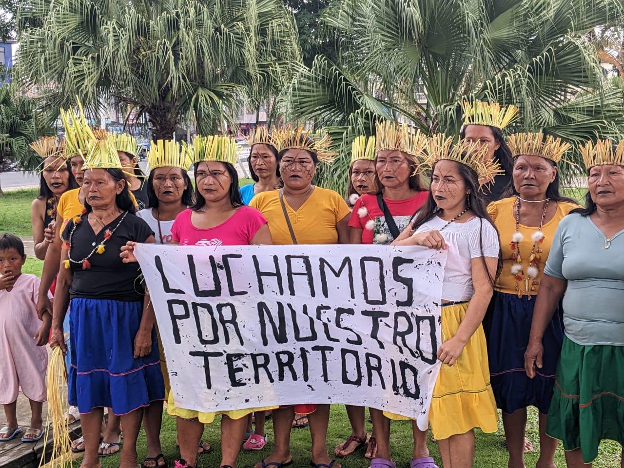 A group of women in Peru wearing headdresses and necklaces from cultures Indigenous to their region stand together with serious facial expressions. They are holding a banner that says "Luchamos por nuestro territorio," which translates to "We fight for our territory."