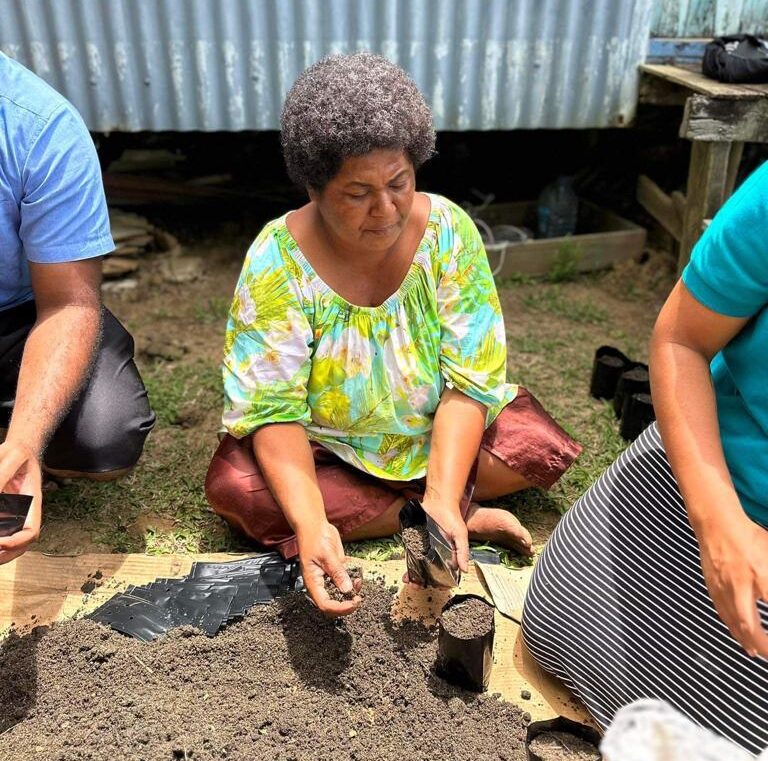 A brown women with an afro sits on grass next to a pile of soil, surrounded by two other people. She is wearing a colorful and bright green and blue shirt patterned with white flowers, and she is focused on scooping soil from the pile into a small container.