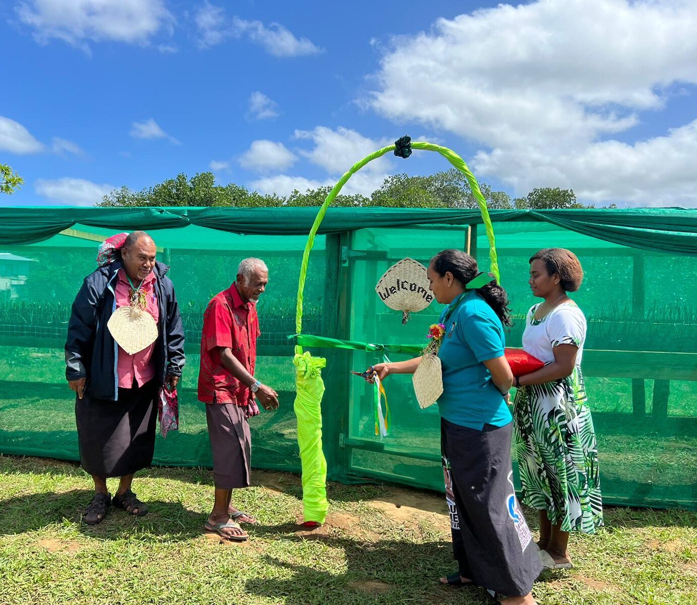 Four brown people stand on either side of a bright green arch, which sits in front of a greenhouse doorway with a "Welcome" sign on it. Someone on the right side of the picture is handing a small object, perhaps a key, to someone on the left side of the picture.