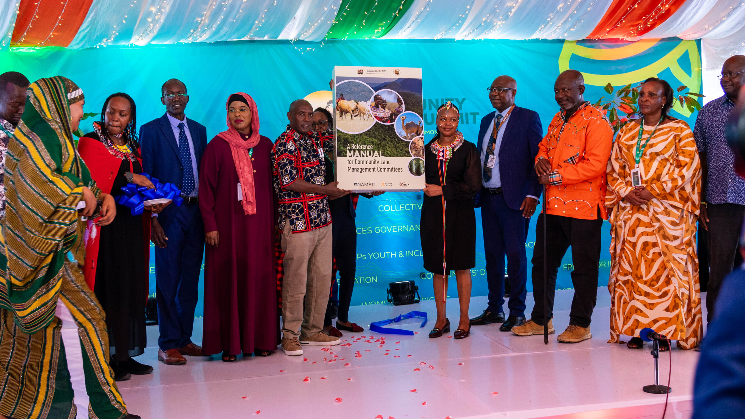 A group of about a dozen Black people with mixed gender presentations, many of them wearing Indigenous clothes and/or accessories, stand together facing the camera. Many of them are turned toward the middle of the group, where two people are holding up a big banner in the shape of a report cover, which has the title "A Reference Manual for Community Land Management Committees" on it.