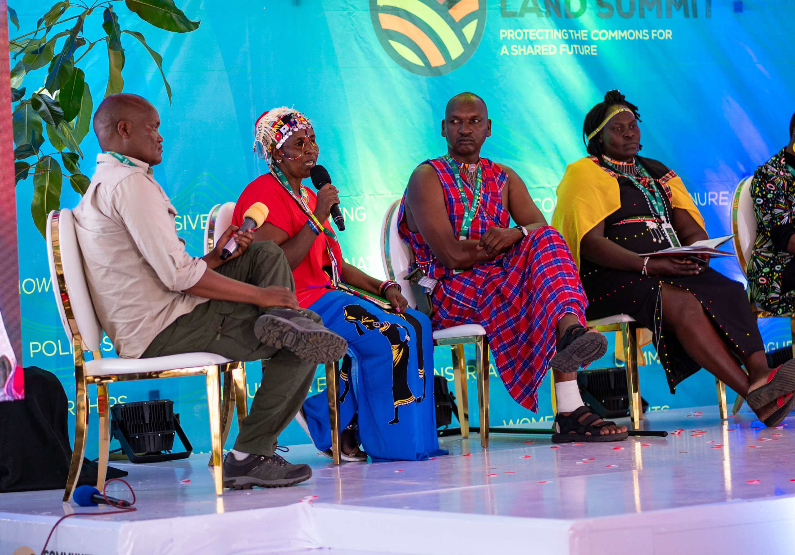 Four Black people, many of them wearing elements of Indigenous clothing and accessories, sit together on a stage. One person is holding a microphone and in the middle of speaking. Behind them is a blue banner that says "Land Summit" Protecting the Commons for a Shared Future."