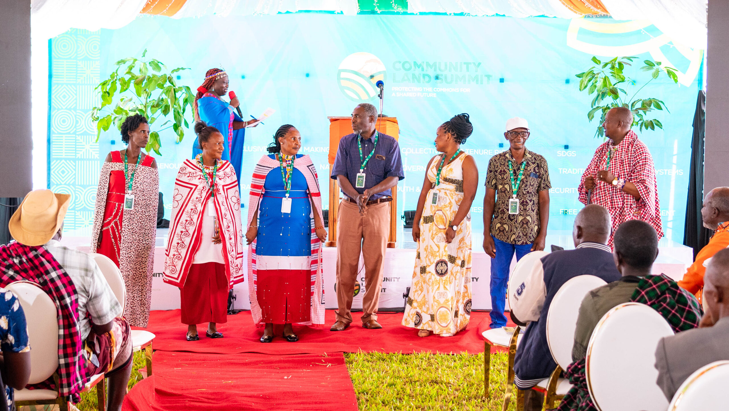 A group of seven Black people, many of them wearing Indigenous clothing and/or accessories, stand side by side facing the camera, which is behind an audience. Behind the group of standing people is a stage, on which is a woman holding a microphone.