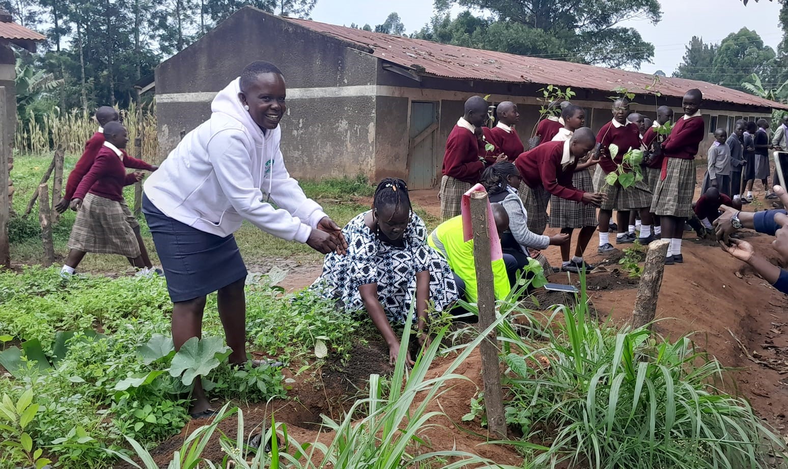 In the foreground, two Black women work together in the soil of a garden full of small plants. One of them is facing the camera and smiling. In the background, many girls in school uniforms hold saplings, seemingly about to plant them.